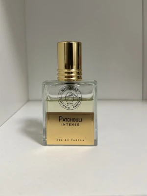 Patchouli Intense Eau de Parfum - Patchouli Intense Eau de Parfum från Nicolai i en elegant, fyrkantig glasflaska med guldfärgat lock. Doften har en varm, kryddig och jordig karaktär med patchouli som huvudnot. Flaskan har en stilren etikett i guld och svart.