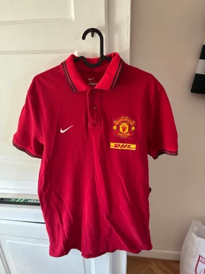 Röd Manchester United pikétröja Nike - Säljer en röd Manchester United pikétröja från Nike med klubbmärke och DHL-logga på bröstet. Tröjan har krage med knappar och svarta detaljer på krage och ärmslut. Perfekt för dig som älskar fotboll och vill representera ditt lag.