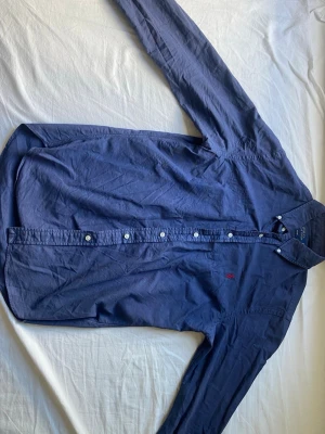 Mörkblå skjorta med knappar - Klassisk mörkblå skjorta med långa ärmar och knappar framtill. Skjortan har en enkel design med liten broderad logga på bröstet. Perfekt till jeans eller chinos för en clean look. Perfekt till allt. 