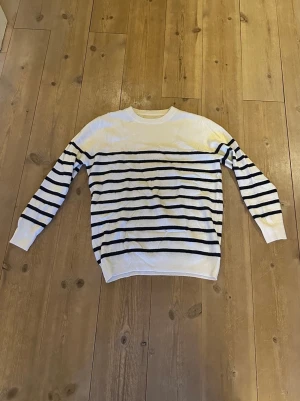 Vit stickad tröja med marinblå ränder - Säljer en vit stickad tröja med marinblå horisontella ränder. Tröjan har rund halsringning, långa ärmar och ribbade muddar vid ärmslut och nederkant. Klassisk och tidlös design som passar till mycket. Helt oanvänd utan defekter. Fraktar inom 1-2 dagar! 