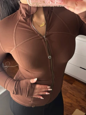 Brun sportjacka med dragkedja - Snygg brun sportjacka med hel dragkedja framtill och hög krage. Jackan har långa ärmar med tumhål och markerade sömmar för en sportig look. Tillverkad i stretchigt funktionsmaterial som passar perfekt till träning eller löpning.