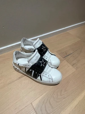 Vita sneakers med svart stjärndetalj - Snygga vita sneakers med bred svart rem över foten, dekorerad med en vit stjärna och nitar runt hälen. Skorna har en låg profil och coola detaljer som ger dem en unik look. Tillverkade i läder med perforeringar på sidan för extra stil. (Dem är lite skadade i bak som visas på bilden men syns ej när dem andvänds💕