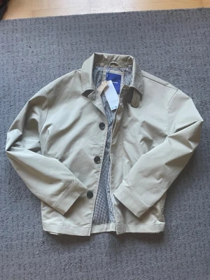 Beige overshirt från Jack & Jones - Snygg beige jacka/overshirt från Jack & Jones med klassisk krage och stora svarta knappar framtill. Insidan har ett rutigt svartvitt foder som ger en cool kontrast. Perfekt för dig som vill ha en stilren och enkel look till vardags.