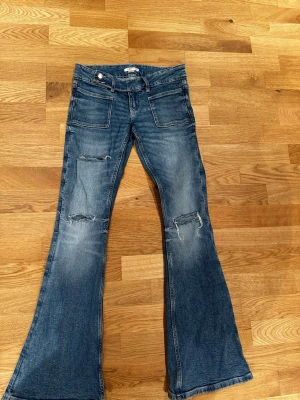 Blå bootcut jeans med slitningar - Säljer ett par blå bootcut jeans med slitningar på båda benen och stora framfickor. Jeansen har en klassisk femficksmodell baktill och är tillverkade i denim. Perfekta för dig som gillar en retro och avslappnad stil.