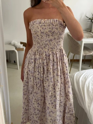 Ljus blommig axelbandslös bandeau klänning - Säljer en söt axelbandslös midiklänning i ljusa toner med småblommigt mönster i lila och beige. Klänningen har smock upptill och ett luftigt, fallande tyg som ger en romantisk känsla. Perfekt för varma dagar och sommarens alla tillfällen.