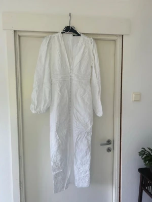 Vit långklänning adoore - En knapp sönder längst ner!!!  Elegant vit långklänning med v-ringning, puffiga långärmar och dekorativa knappar framtill. Klänningen har ett subtilt blommönster och en slits framtill för extra stil. Perfekt för sommarens speciella tillfällen.