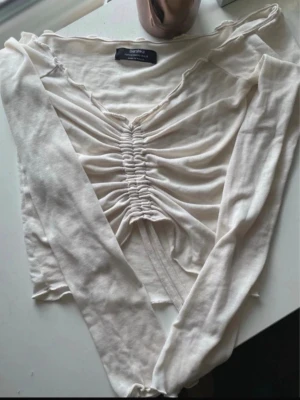Vit långärmad off shoulder topp med snörning - Trendig vit långärmad off shoulder topp med snygg snörning och rynkdetaljer framtill. Toppen har v-ringad hals och är tillverkad i mjukt material som känns skönt mot huden. Perfekt för dig som gillar en modern och stilren look.
