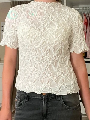 Vit topp med struktur från Vero Moda - Supersöt vit topp från Vero Moda med korta ärmar och unik strukturerad design med små knutar över hela tyget. Toppen har en lätt vågig kant nedtill och är tillverkad i ett tunt, luftigt material som känns skönt mot huden.💕 jag säljer toppen eftersom att det är för liten och därför är en kort på mig💕😅⭐️Kan sänka priset🥰