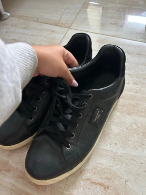 dolce gabbana skor - Säljer ett par svarta låga sneakers i skinn med vita sulor och svarta skosnören. Sidan har en liten svart patch med metallknappar som detalj. Klassisk och stilren modell som passar till det mesta.