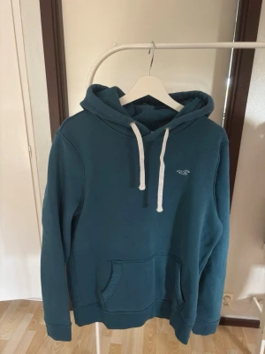 Blå hoodie från Hollister - Mysig blå hoodie från Hollister med vit dragsko och klassisk logga på bröstet. Tillverkad i mjuk fleece för extra komfort. Har stor känguruficka framtill och ribbade muddar vid ärmar och nederkant. Perfekt för chill dagar eller när du vill vara extra bekväm.