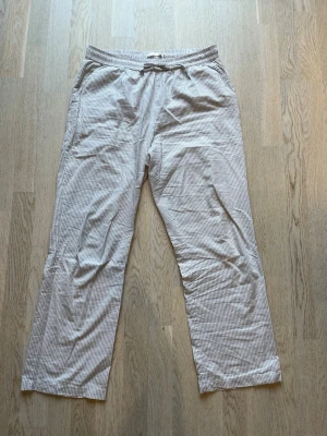 Randiga beige loungewear-byxor - Säljer ett par sköna loungewear-byxor i beige och vitt med tunna vertikala ränder. Byxorna har resår i midjan med dragsko och raka ben. Perfekta för chill dagar hemma eller när du vill vara extra bekväm. Tveka inte på att skriva en fråga eller be om fler bilder.🌸🌸