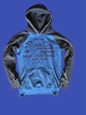 Blå och grå hoodie från Superdry - Snygg hoodie från Superdry i storlek S med blåa med grå ärmar och huva. Stor logga och text på bröstet i vintage-stil. Tröjan har lite slitningar. Fråga gärna om ni undrar något.