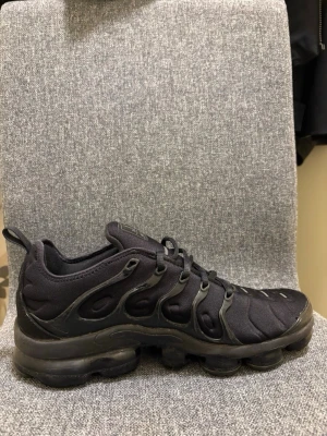 Svarta Nike Air VaporMax Plus sneakers - Snygga helsvarta Nike Air VaporMax Plus sneakers med unik bubbelsula och vågformade detaljer på ovandelen. Skorna har en sportig look med mesh och syntetmaterial, samt diskret Nike-logga på sidan. Perfekta för dig som gillar streetwear och modern stil.