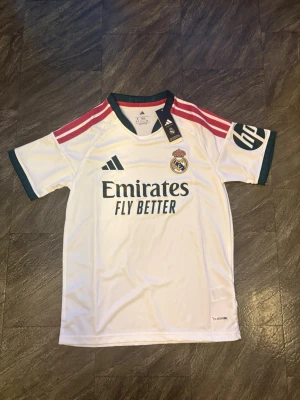 Real Madrid matchtröja Adidas vit 2026/2027 - Snygg vit Real Madrid matchtröja från Adidas med gröna detaljer och rosa ränder på axlarna. Tröjan har klubbmärke, sponsortryck och HP-logga på ärmen. Tillverkad i lätt och ventilerande material, perfekt för fotboll eller träning.