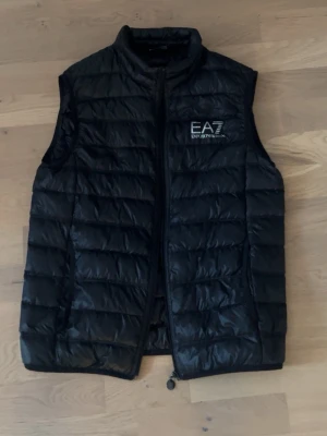 EA7 väst - Snygg svart dunväst från EA7 Emporio Armani med diskret logga på bröstet. Västen har quiltad design, hög krage och dragkedja framtill. Perfekt för lager-på-lager och ger en sportig vibe till din outfit.