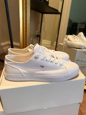 Vita sneakers Tommy Hilfiger - Stilrena vita sneakers från Tommy Hilfiger med lågt skaft och klassisk design. Skorna har vita snören, metallögla vid snörningen och diskret logga på sidan. Tillverkade i tyg. Endast använda en kort stund. Nypris ca 1200kr