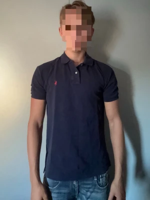 Mörkblå pikétröja från Ralph lauren - Klassisk mörkblå pikétröja med krage och korta ärmar från polo Ralph lauren. Tröjan har två knappar vid halsen och en liten broderad logga på bröstet. Perfekt till jeans eller shorts för en clean och enkel stil. Storlek S