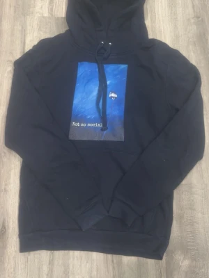 Svart hoodie med blått tryck - Svart hoodie med huva och dragsnören. På bröstet finns ett stort blått tryck med texten 'Not so social' och en liten skylt. Hoodien har magficka och är tillverkad i mjukt material, perfekt för en avslappnad stil.