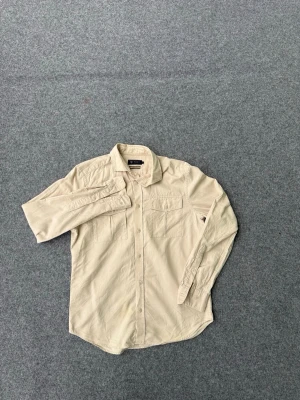 Riley skjorta - Säljer en beige skjorta från Riley i regular fit. Skjortan har två bröstfickor med knappar och klassisk krage. Tillverkad i mjuk bomull och har långa ärmar med knappstängning. Perfekt för en clean och avslappnad stil.