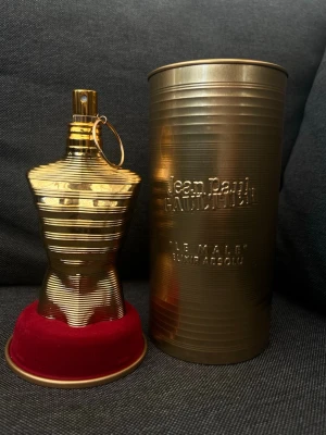 Jean Paul Gaultier  - Unik parfymflaska i form av en manlig torso i guldmetall med horisontella linjer. Flaskan står på en rund röd sammetsliknande bas och kommer med en matchande guldfärgad cylinderformad förpackning. Perfekt för dig som gillar exklusiv design och ikoniska dofter. Parfymen är helt ny använd bara en gång för att känna doften av den. Priset kan sänkas vid snabb affär. 