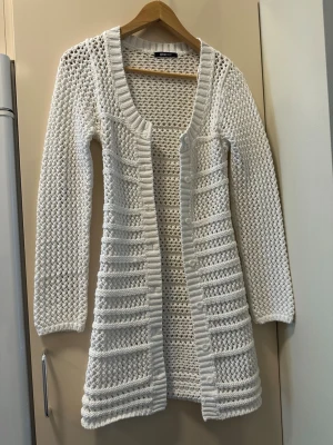 Vit lång stickad kofta Gina Tricot - Säljer en vit lång stickad kofta från Gina Tricot. Koftan har ett öppet hålmönster, ribbade kanter och långa ärmar. Perfekt att slänga över en topp för en chill look. Materialet är mjukt och stretchigt, troligtvis bomull eller bomullsblandning.