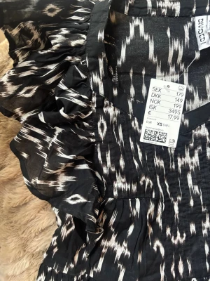 Svartvit mönstrad topp - Svartvit blus med v-ringning och volangdetaljer vid axlarna. Blusen har ett iögonfallande mönster och är tillverkad i ett lätt material, perfekt för varmare dagar. Passformen är normal och modellen har fina rynkade detaljer framtill.