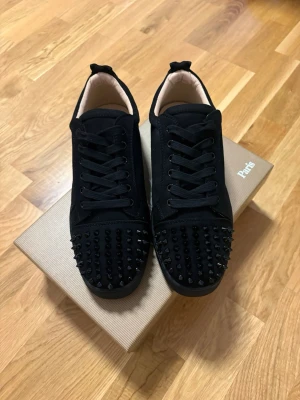 Svarta sneakers med nitar och röd sula - Säljer ett par svarta sneakers från Christian Louboutin med coola svarta nitar på tån och klassisk röd sula. Skorna har snörning, låg profil och är gjorda i mocka och läder. Perfekta för dig som vill sticka ut med din stil. Hör av dig!🤙 