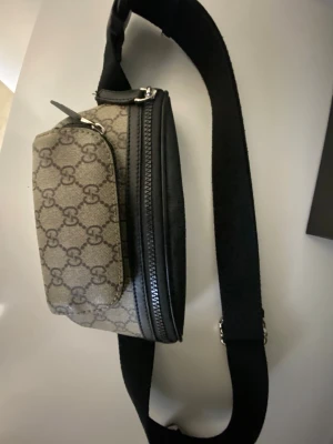Gucci väska - Snygg axelväska från Gucci i beige och brunt med klassiskt GG-monogram. Väskan har svarta detaljer, justerbar axelrem och dragkedja både fram och bak. Perfekt för att bära det viktigaste med stil. Obs relativt ny har endast använt den ett par gånger.