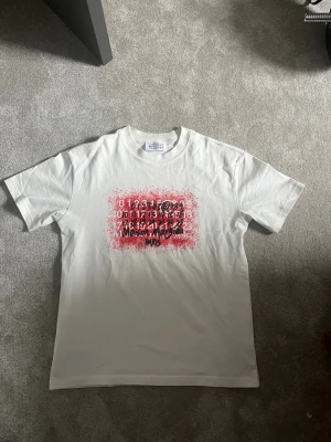 Vit Maison Margiela t-shirt med tryck - Säljer en vit t-shirt från Maison Margiela med ett unikt tryck i rött och svart framtill. T-shirten har korta ärmar och klassisk passform. Perfekt för dig som gillar streetwear och vill sticka ut med cool design.