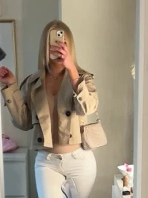 Kort beige zara trenchcoat - Trendig kort trenchcoat i beige med stora svarta knappar och klassisk krage. Jackan har axelklaffar och raka ärmar, samt fickdetaljer framtill. Perfekt för lager-på-lager och ger en snygg siluett till din outfit.