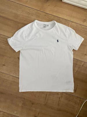 Vit t-shirt från Polo Ralph Lauren - Klassisk vit t-shirt från Polo Ralph Lauren med rund halsringning och den ikoniska mörkblå broderade loggan på bröstet. Tillverkad i mjuk bomull och har en normal passform. Perfekt basplagg till garderoben.