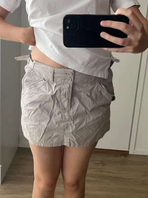 Beige cargo kjol - Säljer en beige kortkjol med dubbla knappar framtill och sidofickor. Kjolen har ett justerbart spänne i midjan för perfekt passform och är gjord i ett lätt bomullsmaterial. Hade behövts strykas till något