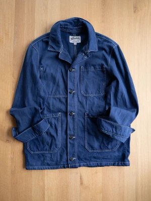 The Stronghold Chore Jacker Blue - Snygg marinblå overshirt med vita kontrastsömmar och tre stora fickor framtill. Jackan har klassisk krage, knäppning med knappar och är tillverkad i bomull. Perfekt för lager på lager och enkel att styla till olika outfits.