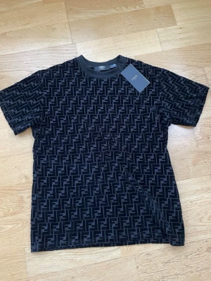 Svart mönstrad t-shirt från Fendi - Snygg svart t-shirt från Fendi med det klassiska FF-mönstret över hela plagget. T-shirten har rund halsringning och korta ärmar. Perfekt för dig som vill ha en statement piece i garderoben.