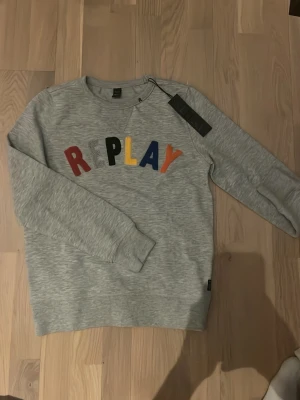 Grå sweatshirt från Replay - En sprilansny snygg grå sweatshirt från Replay med färgglada bokstäver på bröstet. Tröjan har rund halsringning, långa ärmar och ribbade muddar. Perfekt för en avslappnad och trendig stil.