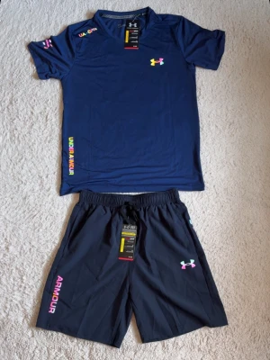 Blått träningsset från Under Armour - Snyggt träningsset från Under Armour med en blå t-shirt och matchande shorts. T-shirten har färgglada logotyper och tryck, rund hals och korta ärmar. Shortsen har elastisk midja och färgglad logga på benet. Perfekt för gym eller löpning.