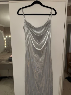 Lång silverklänning med smala axelband - Snygg långklänning i glansigt silvermaterial med smala axelband och draperad urringning. Klänningen har en elegant och figurnära passform