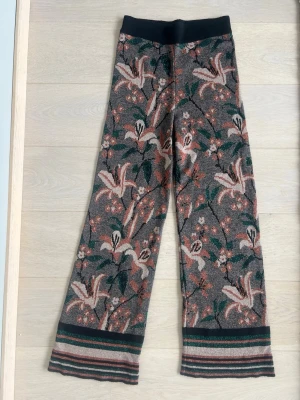 Vida stickade byxor med blommor - Unika, vida stickade byxor med blommigt mönster i grönt, rosa, beige och svart. Byxorna har lätt elastisk midja. För korta för mig som är 179. Är Italiensk storlek M, men passar mig i midjan som är XS eftersom det är så mjuka och elastiska i midjan. Endast använda några fåtal gånger.