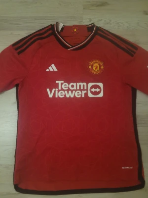 Manchester United matchtröja Adidas - Säljer en röd Manchester United matchtröja från Adidas med svarta detaljer och klubbmärke på bröstet. Tröjan har TeamViewer-tryck på framsidan och klassiska Adidas-ränder på axlarna. Tillverkad i lätt funktionsmaterial, perfekt för fotbollsträning.