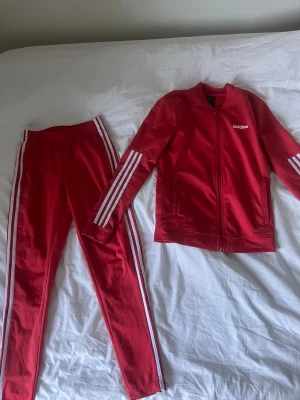 Rött Adidas träningsset med vita ränder - Säljer ett rött träningsset från Adidas med klassiska vita ränder längs ärmar och ben. Setet består av en zip-jacka och matchande byxor i stretchigt material. Perfekt för träning eller chill. Adidas-logga på både jacka och byxa.