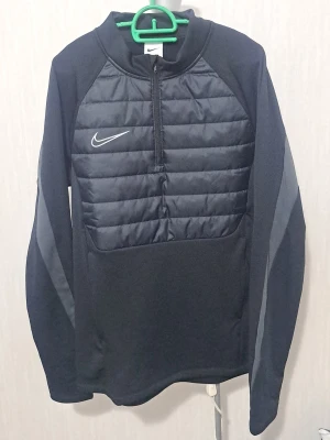 Nike oanvänd - Svart träningsjacka från Nike med quiltad frontpanel och halv dragkedja. Bara testad! Jackan har långa ärmar med gråa detaljer längs sidorna och hög krage. Tillverkad i syntetmaterial som passar perfekt för löpning eller utomhusträning när det är kallare.