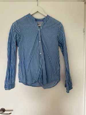 Blåvit randig skjorta med knappar - Snygg blå skjorta med vita vertikala ränder och knappar framtill. Skjortan har lång ärm och en avslappnad passform, perfekt för en stilren vardagslook. Tillverkad i ett lätt och bekvämt bomullsmaterial.