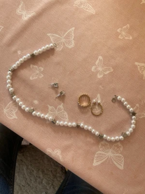 Pärlsmyckeset med ring och örhängen - Säljer ett smyckeset med ett pärlhalsband, ett par guldiga ringörhängen, ett par små silvriga örhängen och en justerbar ring i guldton med två vita pärlor. Materialen är metall och konstpärlor. Perfekt för dig som gillar klassiska och eleganta accessoarer. Är helt oanvända 