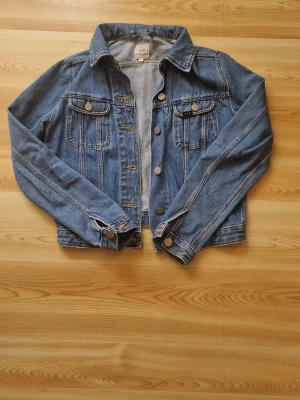 Blå jeansjacka från Lee - Klassisk blå jeansjacka från Lee med två bröstfickor med lock och knappar, samt knappstängning framtill. Snygga sömmdetaljer och justerbara ärmslut. Perfekt att slänga över en hoodie eller t-shirt för en avslappnad look, Inga defekter och i mycket bra skick. Storlek s, passar s/xs.💗