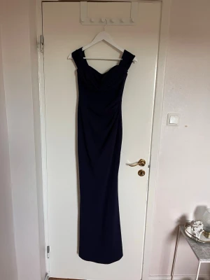 Mörkblå långklänning med breda axelband - Elegant mörkblå långklänning med breda axelband och figurnära passform. Klänningen har en markerad byst och rak siluett som ger en stilren look. Perfekt för speciella tillfällen när du vill känna dig extra snygg.
