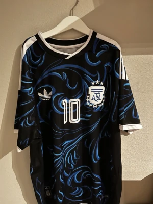 Argentina Messi tröja svart/blå - Säljer en svart och blå Argentina fotbollströja med nummer 10 och Messi på ryggen. Tröjan har vita detaljer, AFA-logga och Adidas-märke på bröstet. Snyggt mönster i blått över hela tröjan och vita ränder på axlarna. Perfekt för fotbollsfans!