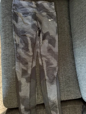 Svarta Nike leggings med mönster - Säljer ett par svarta Nike leggings med vitt rutmönster och meshdetaljer längs benen. De har en tight passform och Nike-logga på höften. Perfekta för träning eller chill dagar. Materialet är stretchigt och andas bra.