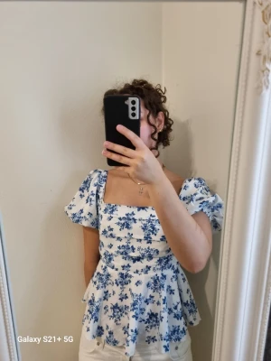 Vit blommig blus med puffärmar - Supersöt blus med blått blommönster på vit botten. Modellen har puffiga korta ärmar, fyrkantig ringning och lätt utsvängd midja som ger en romantisk vibe. Perfekt för dig som vill ha en feminin och trendig topp till sommaren.