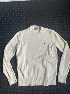 Beige stickad tröja med brodyr - Klassisk beige stickad tröja med rund hals och ribbade muddar. Tröjan har en liten mörkblå broderad detalj på bröstet. Perfekt basplagg för lager på lager och enkel att matcha med olika stilar.