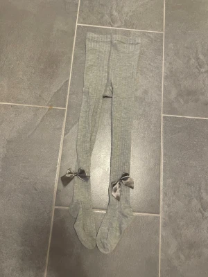 Grå ribbade strumpbyxor med rosetter - Säljer ett par ljusgrå ribbade strumpbyxor med dekorativa satinrosetter vid anklarna. Strumpbyxan är stretchiga och mjuka, perfekta för att ge en söt touch till din outfit. Materialet känns bekvämt och passar bra till olika stilar.